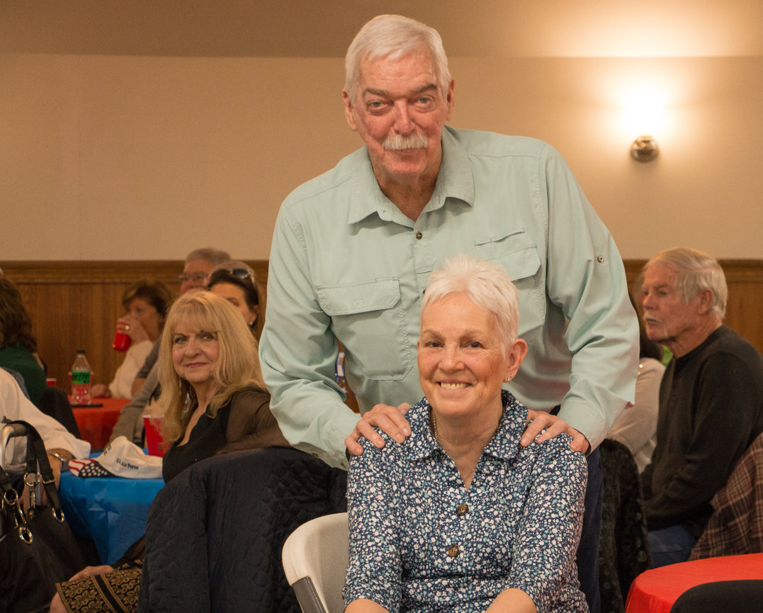Older couple smiling, with the man standing and woman seated in a crowded, warmly lit room.