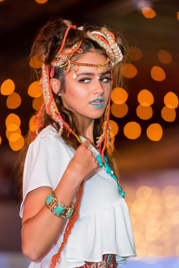 Fashion model with intricate braided and dreadlock hairstyle adorned with colorful yarn and beads, wearing turquoise jewelry, a white ruffled top, and blue lipstick, walking on a runway with warm bokeh lights in the background