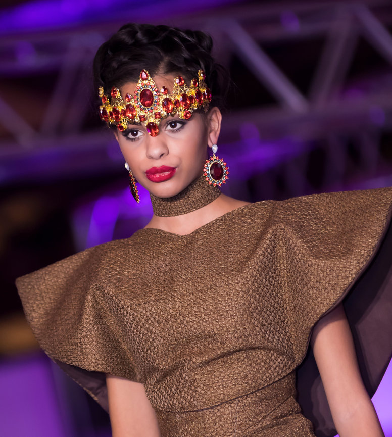Fashion model wearing a textured brown structured dress with dramatic sleeves, a matching high neck, and an ornate gold and red gemstone headpiece and earrings, walking on a runway with a blurred purple-lit background.