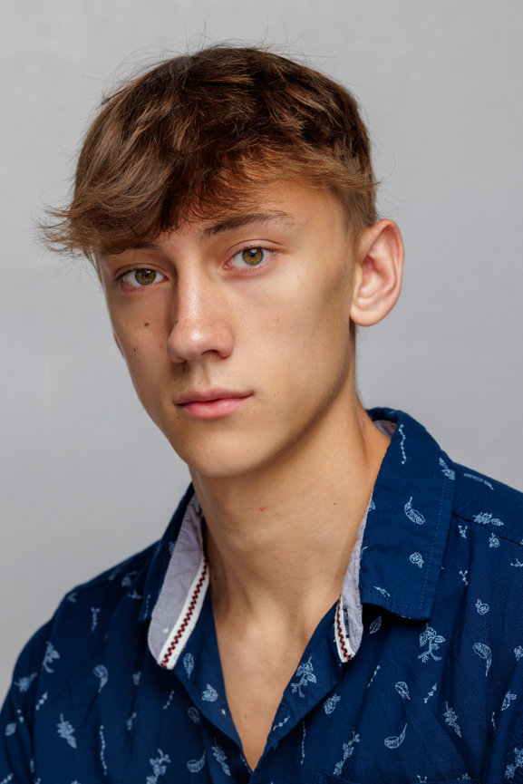 Portrait of a young man with short brown hair, wearing a blue patterned shirt against a light background, taken by Patrick Handy
