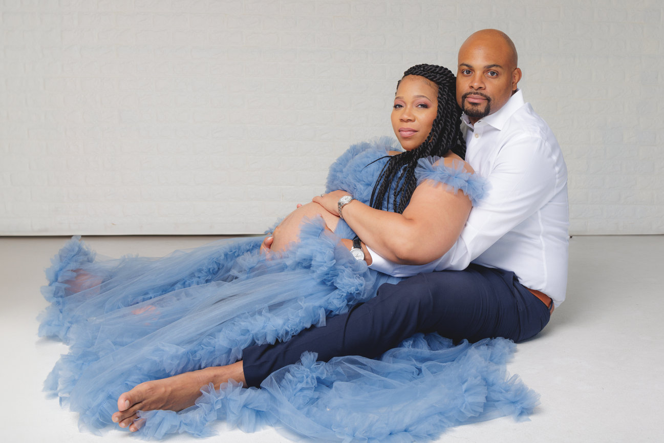 Couple seated together in an elegant pose, featuring the pregnant woman in a blue tulle dress and the man in a white shirt.