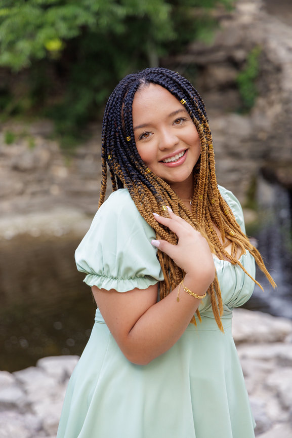 Graduate with braided hair smiles while wearing a light green dress, standing by a natural water feature Prairie Creek Park in Richardson, Texas.
