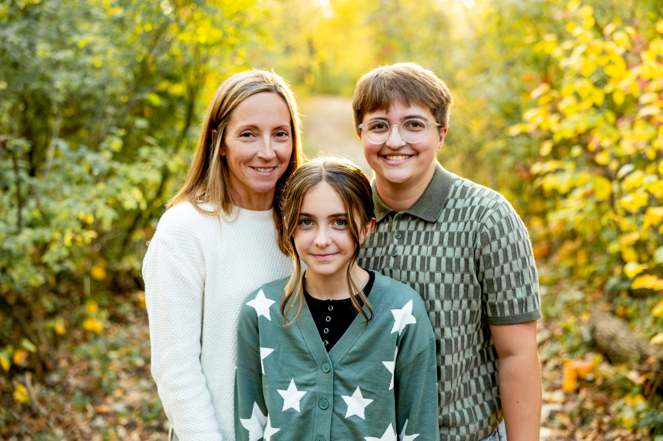Three people pose outdoors on a leafy path in autumn, surrounded by vibrant yellow and green foliage.