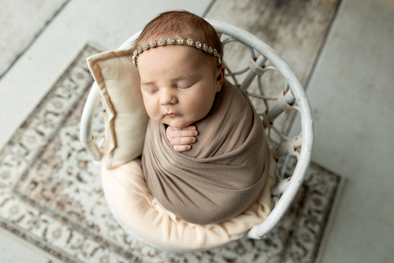 Baby wrapped in taupe blanket, sleeping in a circular basket with a floral headband.
