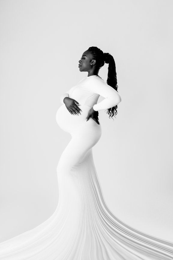 Pregnant woman in flowing white gown, hands on back, side profile, with neutral background.