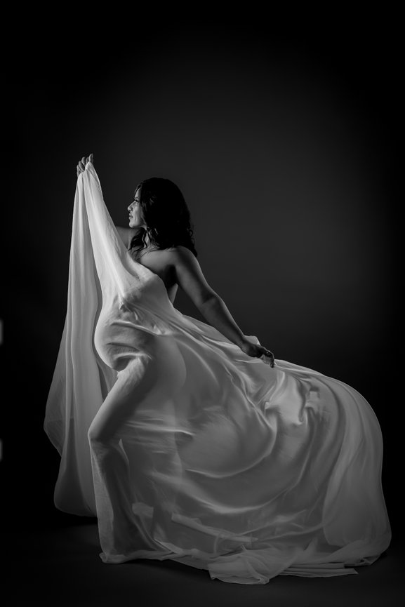 Pregnant woman elegantly poses with flowing fabric in a dramatic black and white setting.