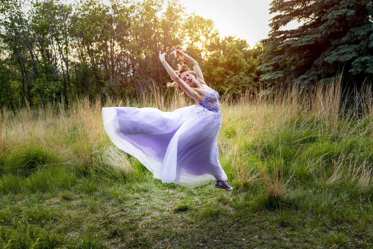 Person in a flowing lavender dress dancing in a grassy field with trees and a setting sun.