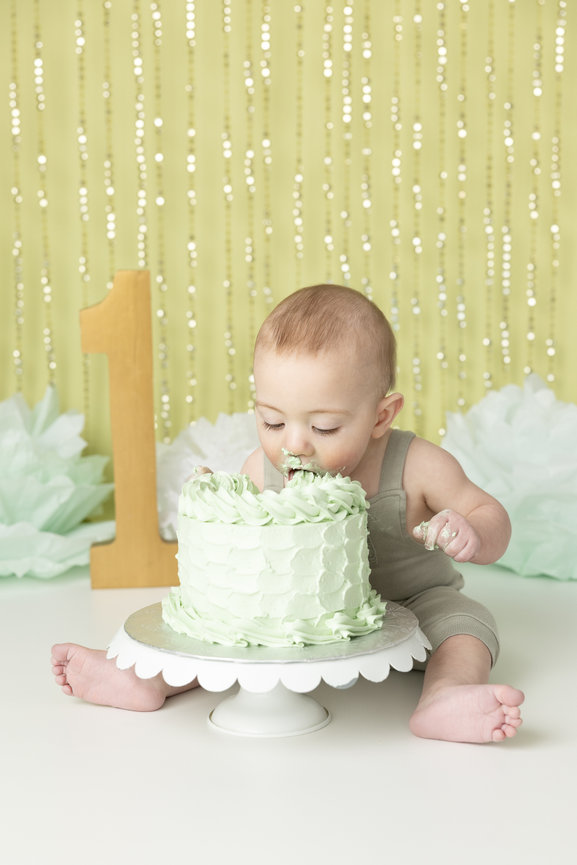 Baby in a romper enjoying a green cake, sitting on the floor, with decorative backdrop and a large number one.