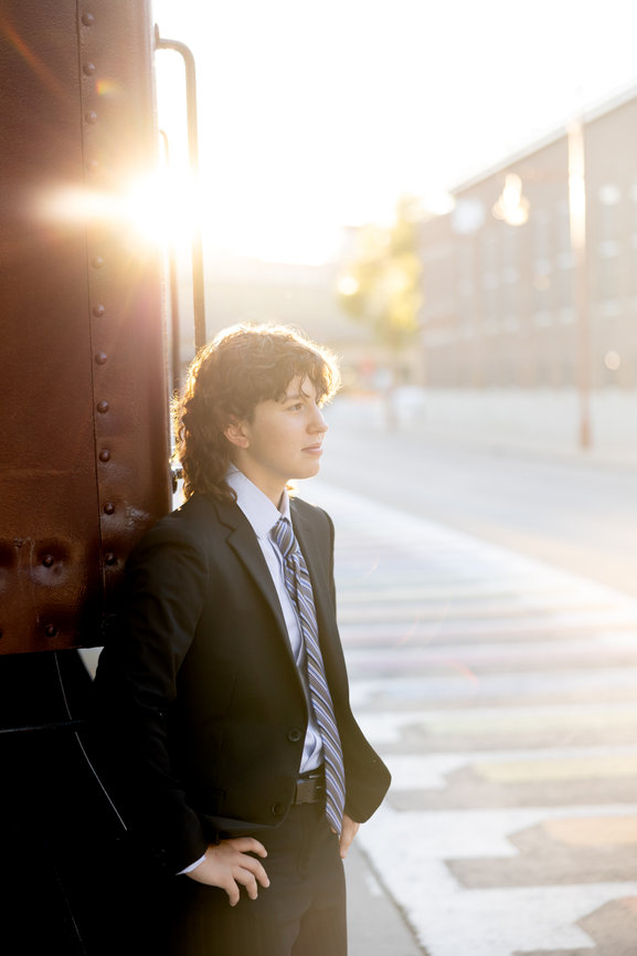 Graduate in a suit leaning against a train car in bright sunlight