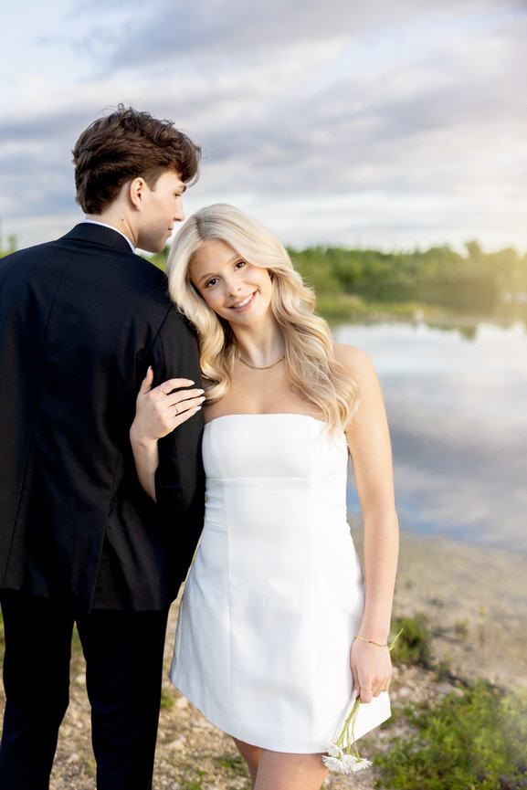 Young couple posing by a lake, woman in white dress holding flowers and man in black suit