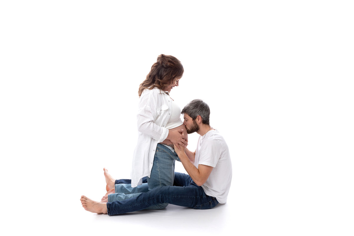 A young couple in white shirts and blue jeans posing on a white backdrop on the ground for their maternity session.