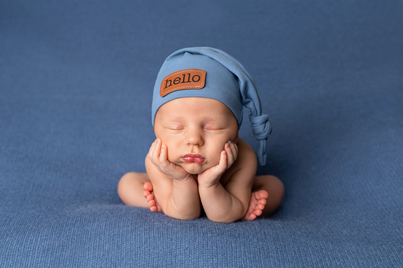 Sleeping baby wearing a blue hat with a hello tag, resting on a blue blanket during his newborn session at the studio of Angi Randall Photography.