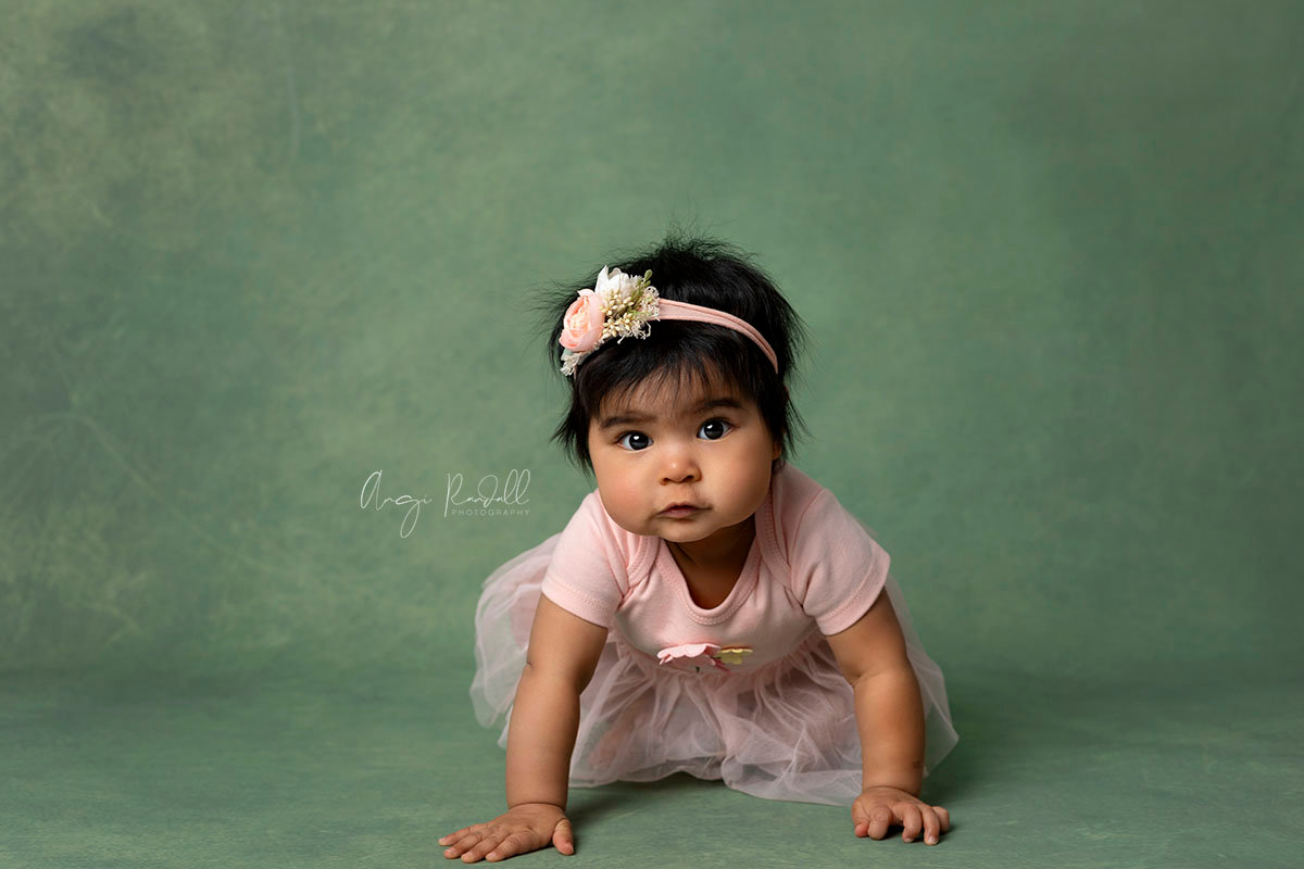 baby girl in a peach dress with matching headband crawling on a green backdrop. eau claire baby photographer.