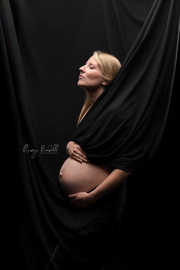 Pregnant woman posing in black draped fabric against a dark studio background in Augusta, Wisconsin at the studio of Angi Randall Photography.