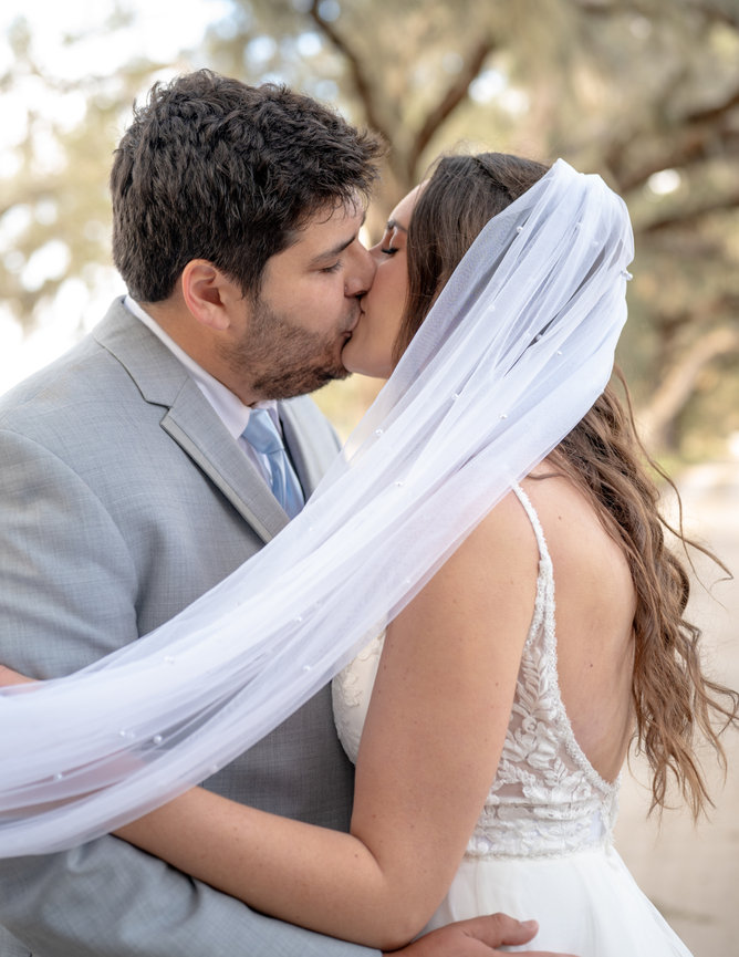Bride and groom kissing under a flowing veil outdoors.