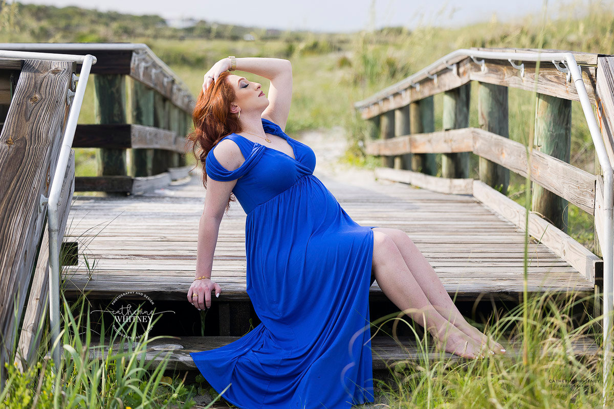 A pregnant woman in a blue dress sits on a wooden bridge in a grassy field, striking a relaxed pose at Hanna Park Beach during a maternity photography session with Catherine Whitney Photography in Jacksonville Florida