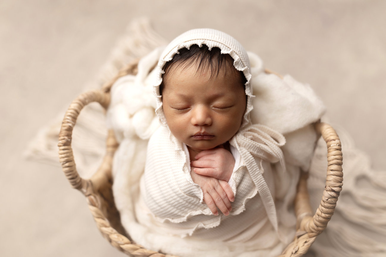 Bennington photographer captures newborn baby asleep in a basket, wrapped in a white blanket and wearing a matching hat.