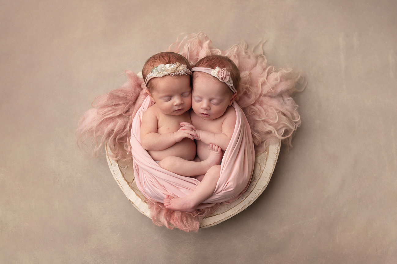 Two sleeping newborns, twins, wrapped in soft pink fabric resting in a round basket.