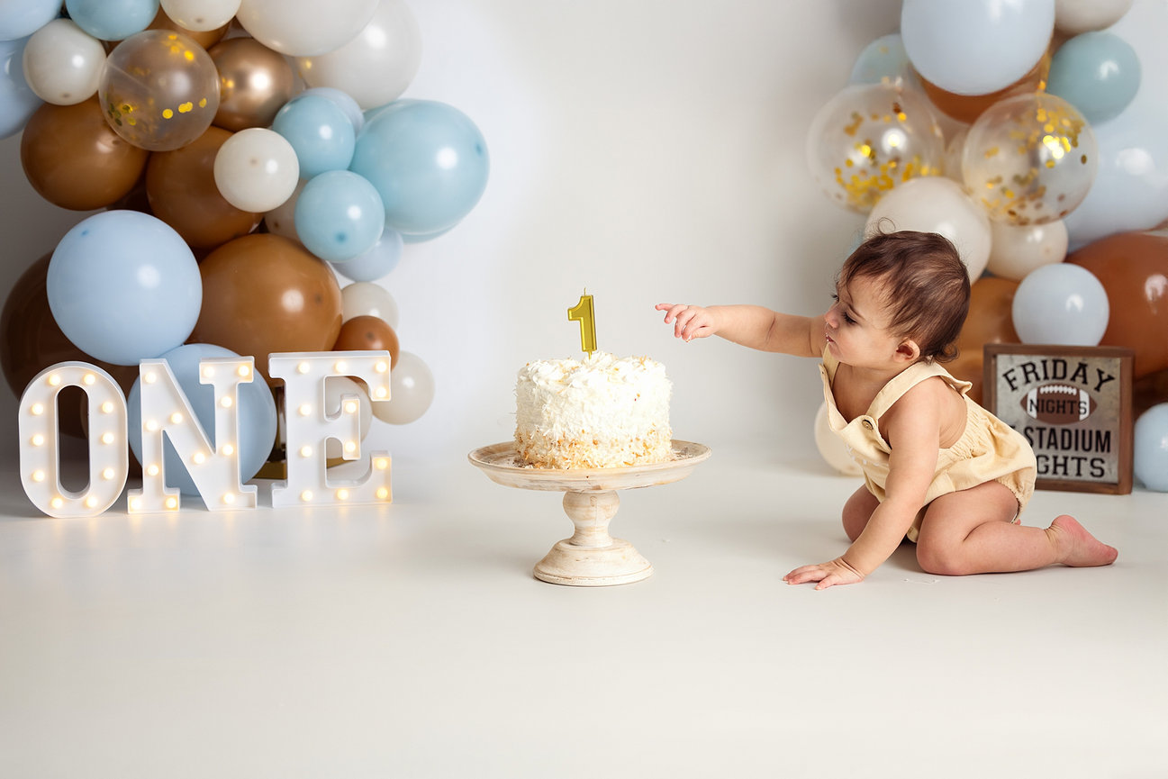 A baby girl in a pink dress sits before a beautifully decorated cake, her expression full of delight during her cake smash photo session with Catherine Whitney Photography