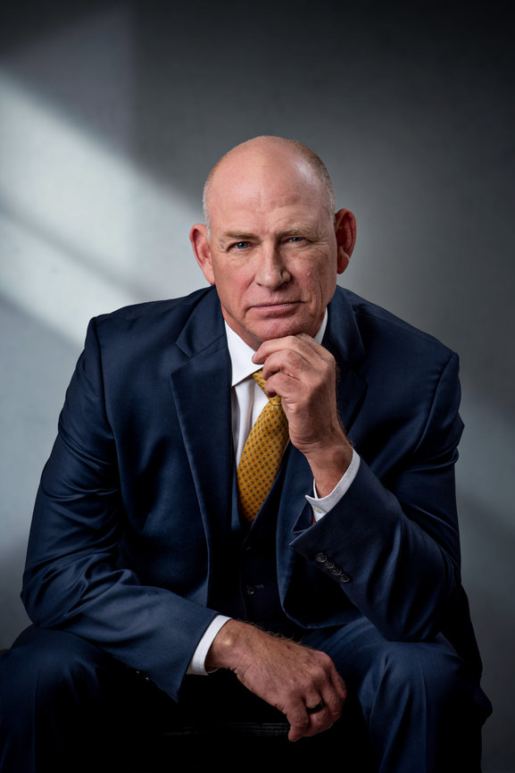 An older man in a suit sitting leaning forward with his hand under his chin.