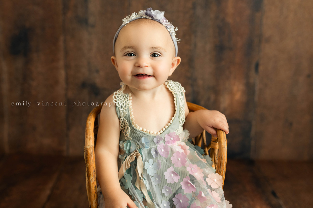 smiling baby girl in floral dress and headband smiling and sitting, posing with neutral background