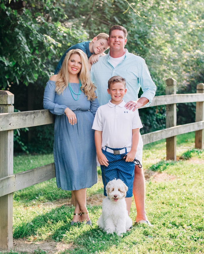 A family of four with a dog poses by a wooden fence in a sunny park. The parents stand behind two boys, one of whom is leaning on his dad.