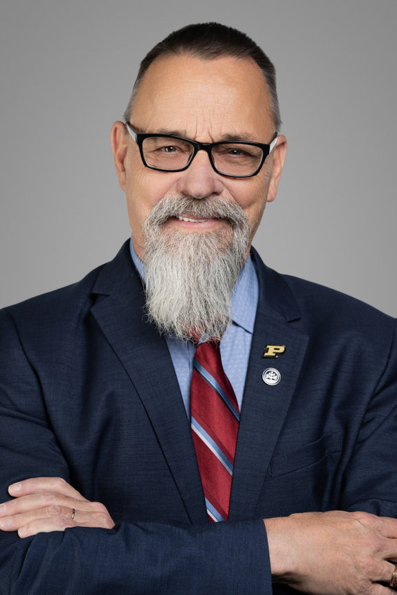 Professional headshot of a man with facial hair, wearing glasses, a suit, and a tie, smiling confidently.