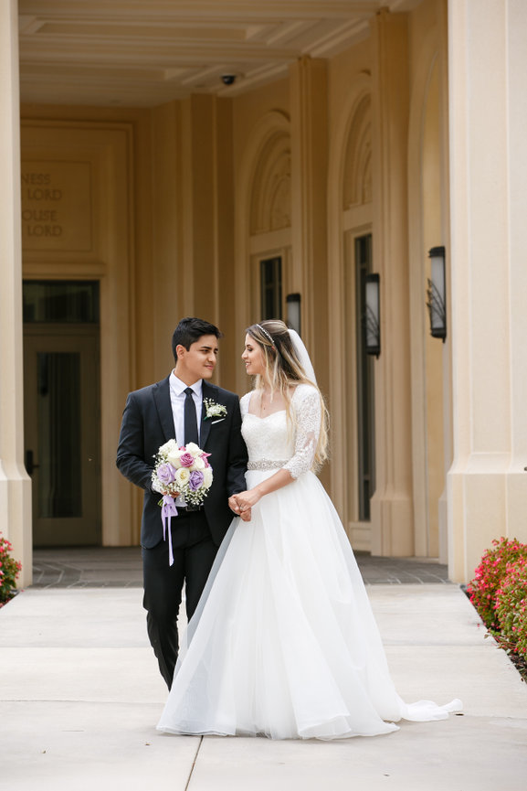 A bride and groom holding hands, gazing at each other, standing outside an elegant building.
