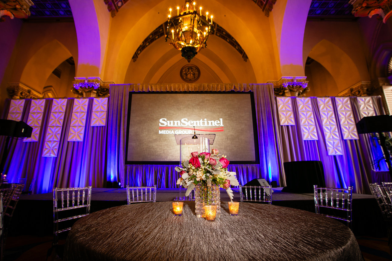 Elegant banquet hall stage with Sun Sentinel Media Group screen, floral centerpiece, and purple lighting