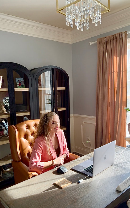 Woman in pink jacket sits at a desk using a laptop in a bright home office headshot photographer for women Maryland.
