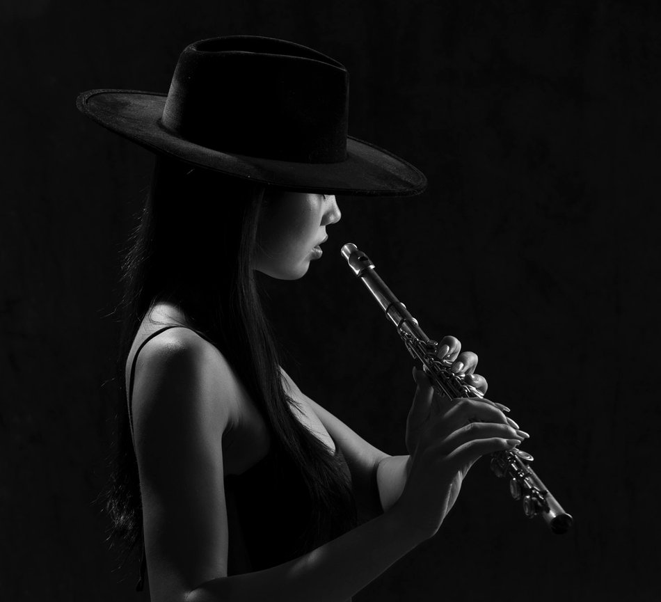 Monochrome portrait of a woman in a wide-brim hat playing a flute on a dark background with dramatic lighting