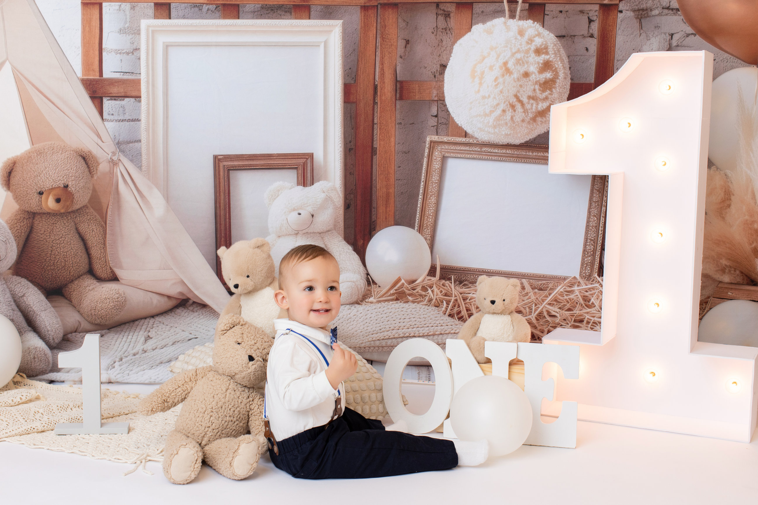 Baby in blue suspenders seated among teddy bears, vintage suitcases and wooden ONE letters