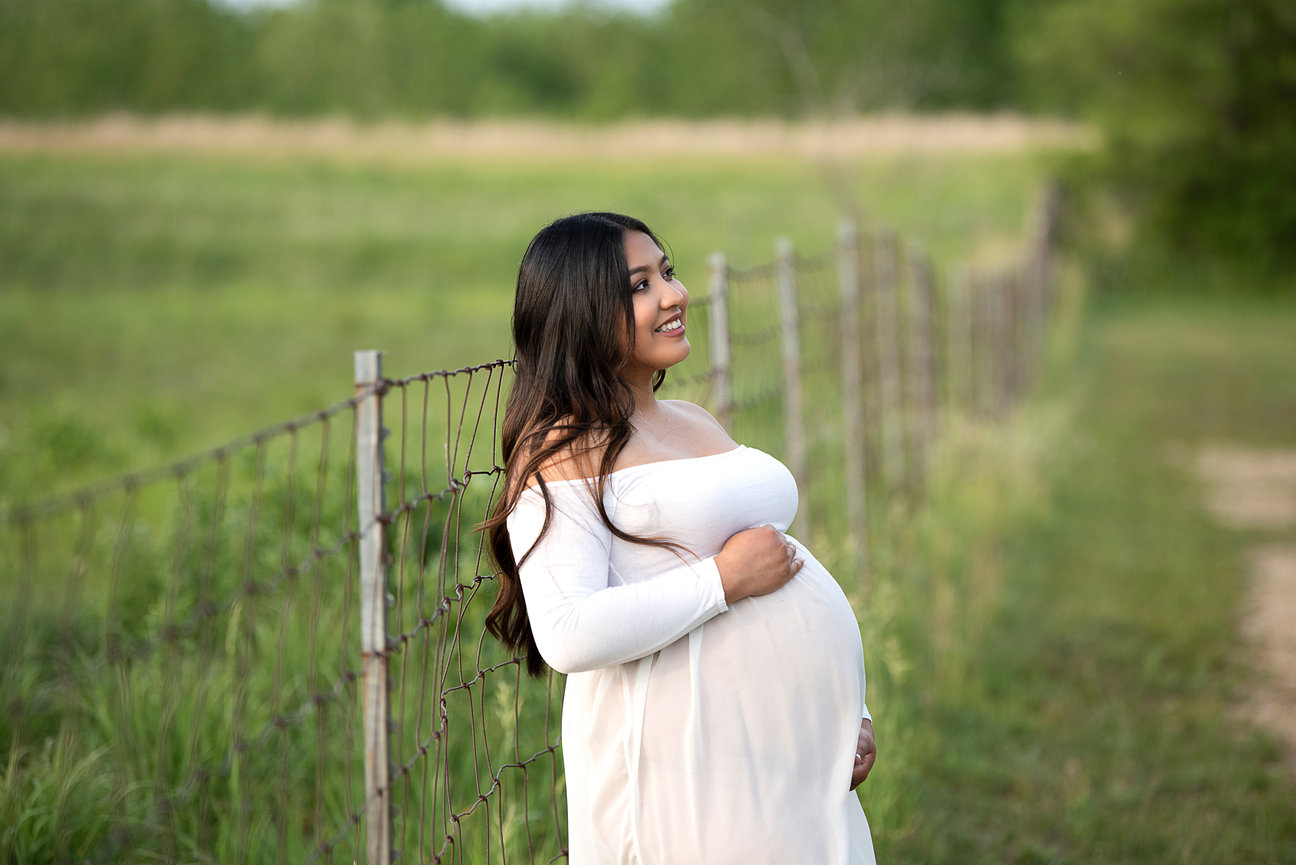 Clad in a white dress, the radiant, expectant woman leans against a wire fence in a lush green field. Her smile reflects pure joy as she lovingly cradles her belly, embracing the beauty of maternity surrounded by trees and grass.