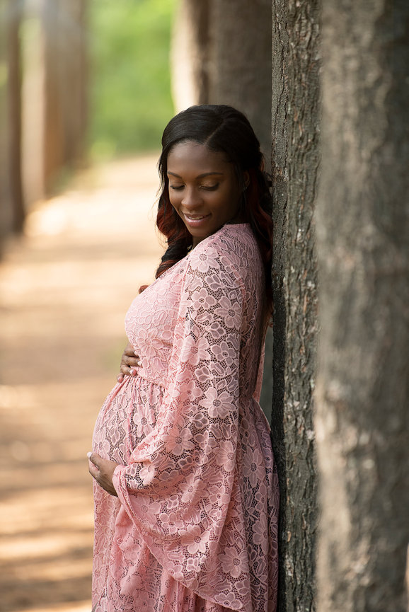 A pregnant woman in a pink lace dress stands outdoors, leaning against a tree with her hand gently on her belly. She smiles softly, surrounded by a blurred background of trees and a sunlit path.