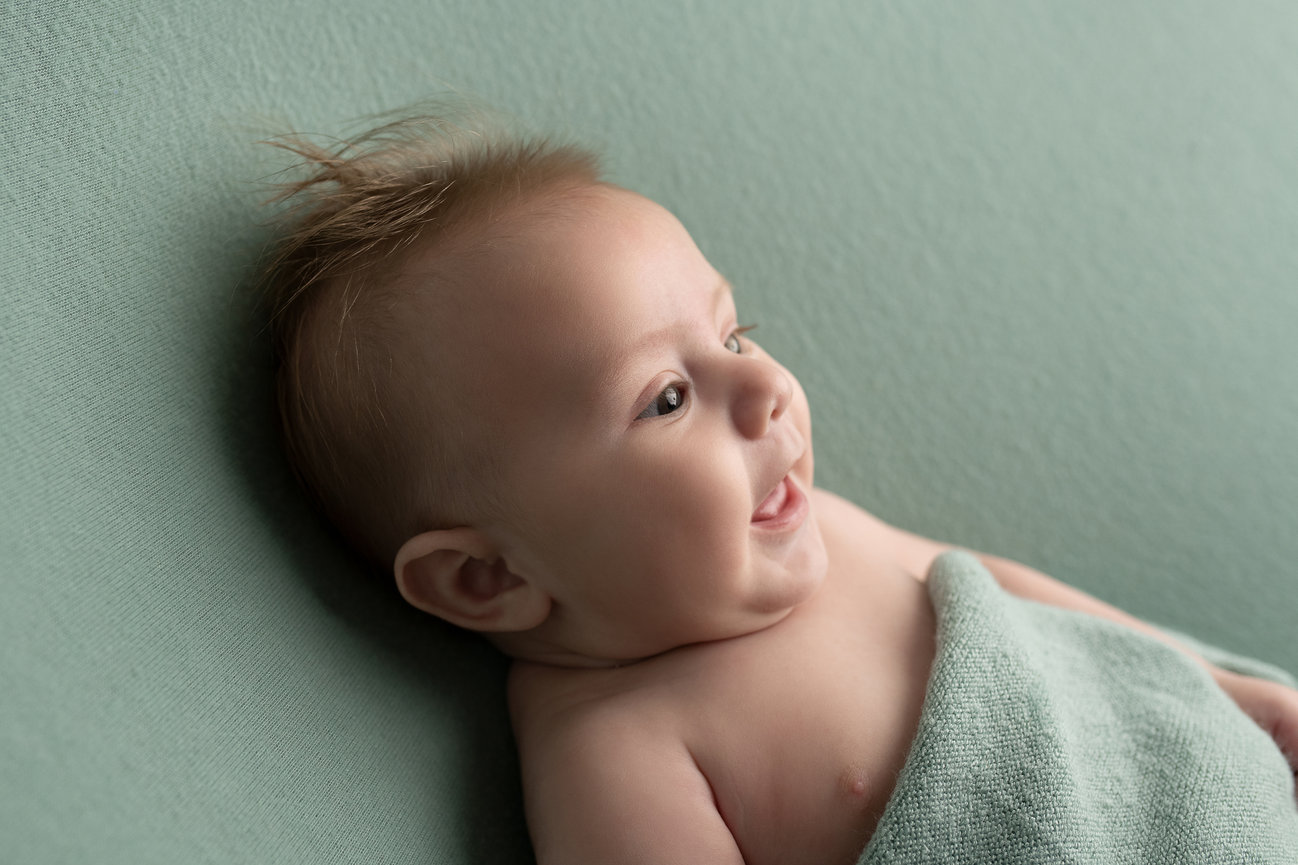 A smiling baby with fine hair lies wrapped in a soft, light green blanket against a matching green background, reminiscent of the peaceful suburbs of Chicago, looking to the right.