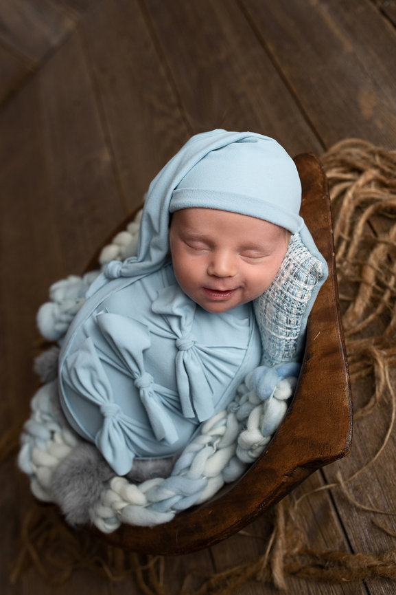 A newborn baby peacefully sleeps in a wooden bowl, wrapped in a light blue swaddle with large bows and matching hat. The cozy setup, reminiscent of West Chicagos charming rustic style, includes soft knitted blankets and a rustic wooden background.