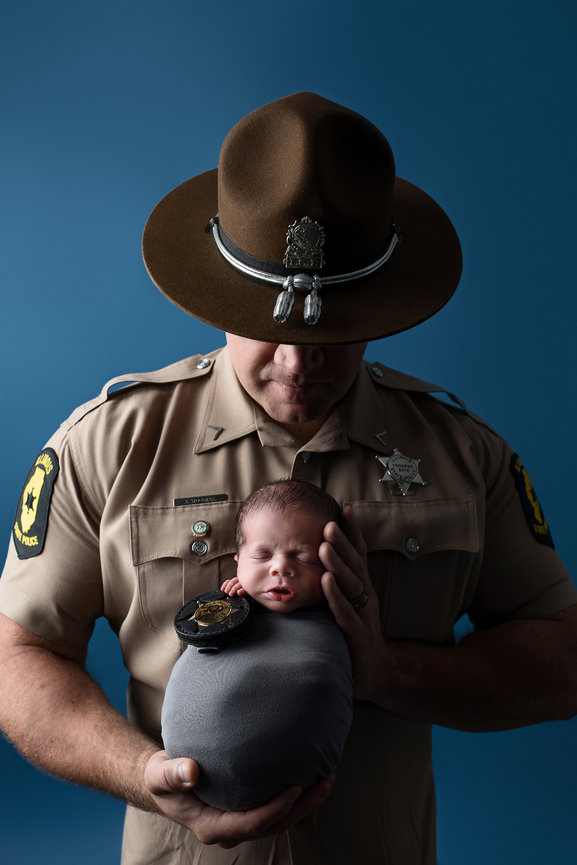 A uniformed officer from Bartlett, Illinois, gently holds a sleeping newborn wrapped in a gray blanket. The baby rests its head on the officers hand, with a badge placed on the blanket. The background is a solid blue.