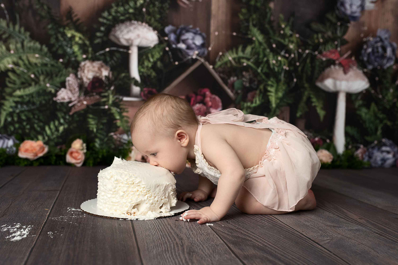 A baby in a pink outfit leans forward on a wooden floor for an exciting 1st birthday cake smash, taking a bite of white frosted cake. The backdrop is adorned with lush greenery, vibrant flowers, and large decorative mushrooms.