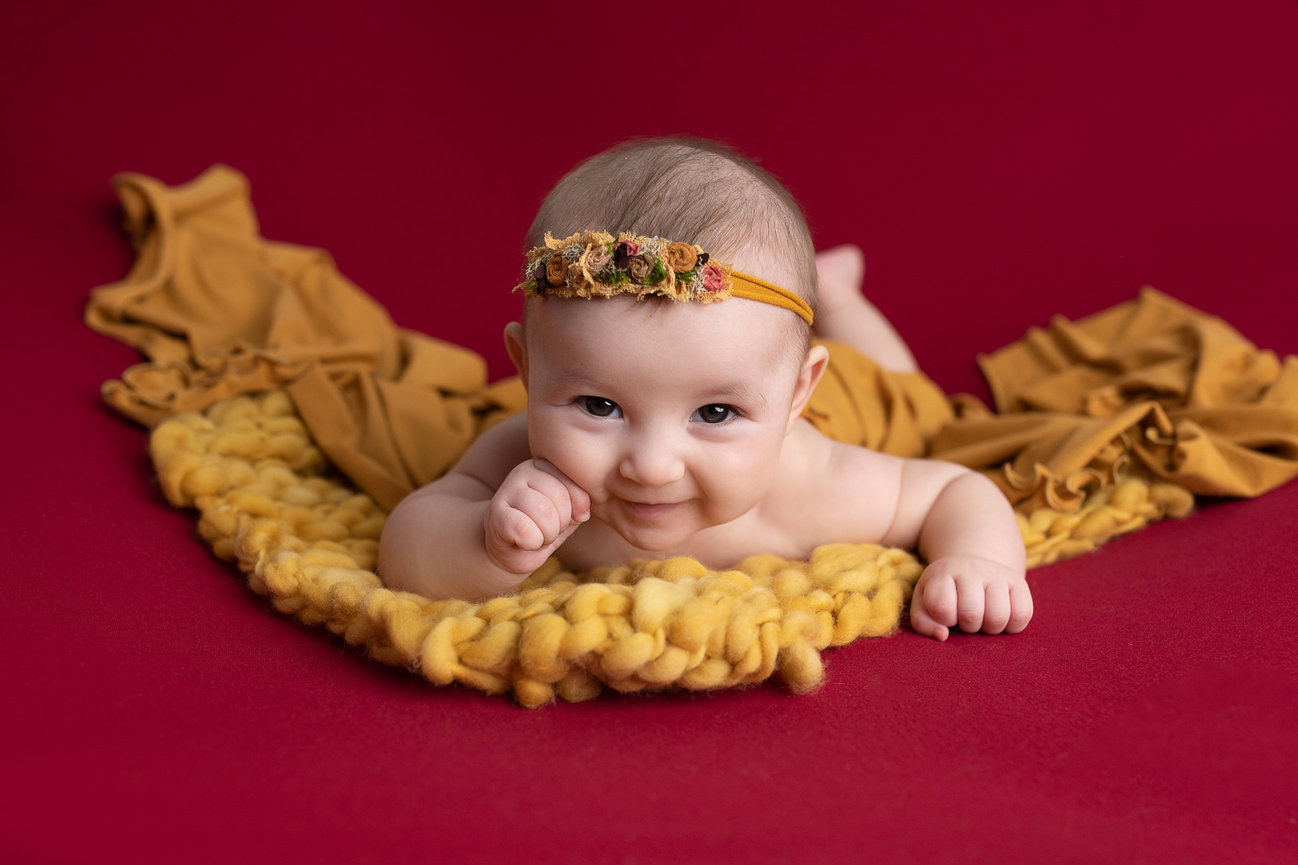 A baby lies on a soft, mustard-colored blanket, set against a vibrant red backdrop. Dressed with a floral headband, the little one poses playfully for the photo shoot, resting on their stomach with one hand near their mouth and eyes full of curiosity