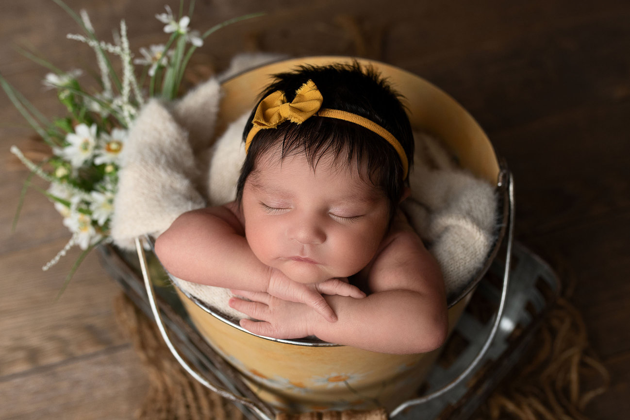 A sleeping baby with dark hair rests peacefully in a bucket, cushioned by a soft blanket. The little one wears a yellow headband with a bow. White flowers add charm to the rustic setting of wooden textures, near Geneva, Illinois.