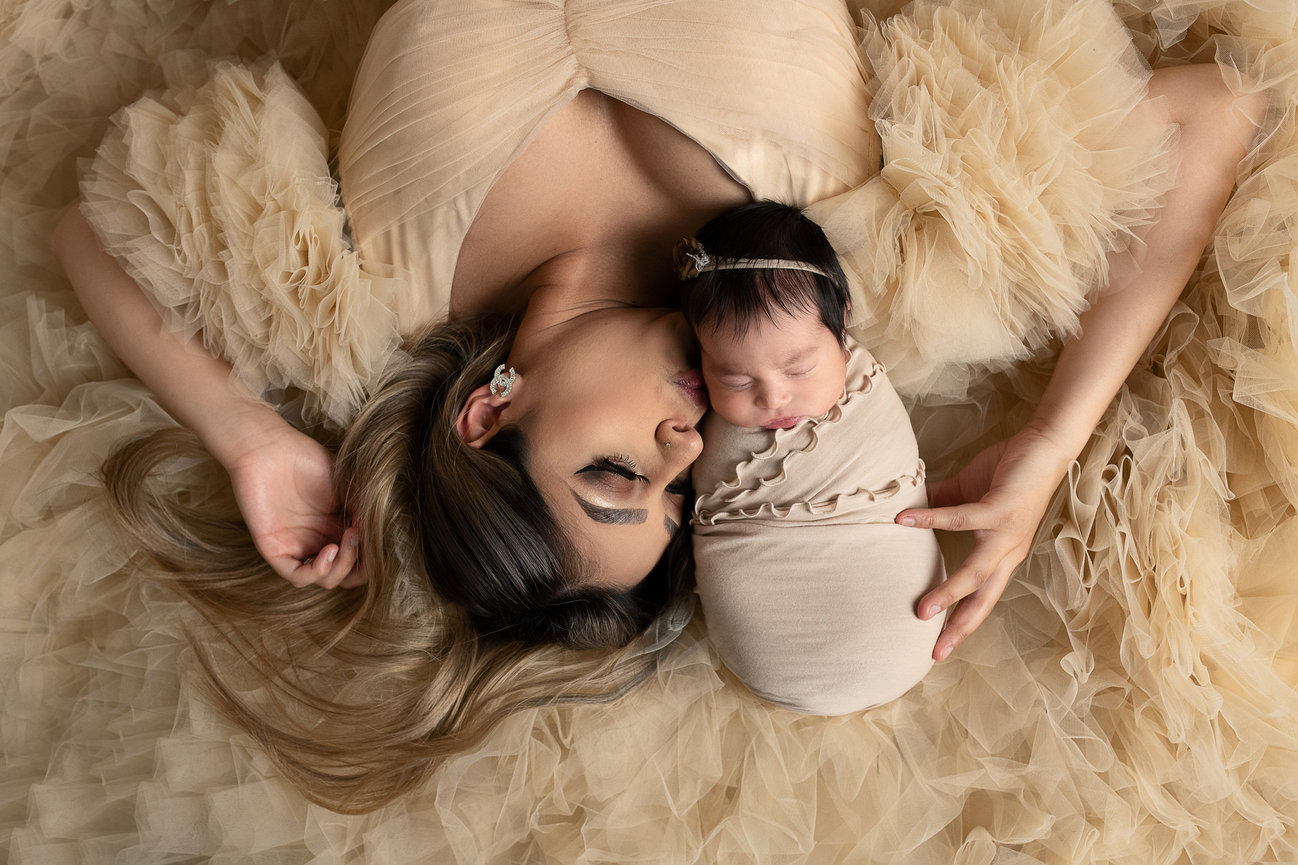A woman lying on her back in Pingree Grove, Illinois, eyes closed, kisses a sleeping newborn wrapped in a beige blanket. Both are enveloped by soft, ruffled beige fabric, creating a serene and tender atmosphere