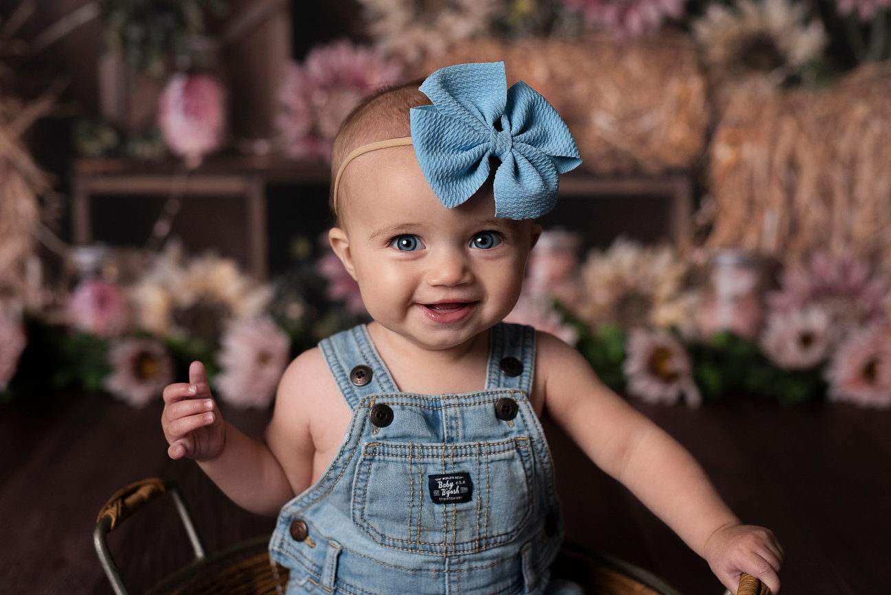 In a portrait studio setting, a baby with blue eyes beams while sitting in a basket, donned in denim overalls and a large blue bow headband. The background offers flowers, hay bales, and soft lighting that enhance the charming scene.