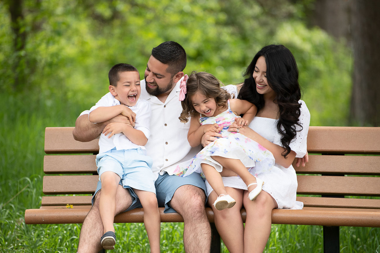 A family of four sits joyfully on a park bench in Cary, Illinois. Two adults and two children laugh and play together, dressed in summer clothes. The scene is set against a backdrop of lush green trees and grass, capturing the essence of a sunny day.