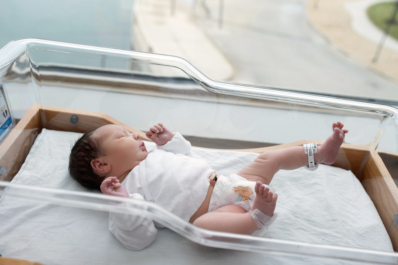 A newborn baby lies in a hospital bassinet at Huntley Hospital, dressed in a white outfit and diaper. Identification bands adorn the tiny wrist and ankle. The bassinet is cozily positioned near a large window, offering a view of the world outside.