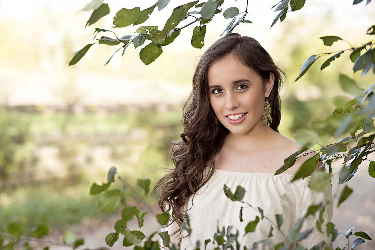 A woman with long brown hair smiles, surrounded by green leaves. She wears a white off-shoulder top and earrings, captured in a high school photography style. The blurred background suggests a natural outdoor setting.
