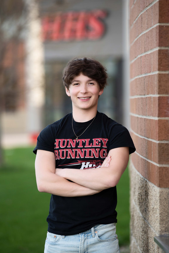 A teenager with short brown hair, wearing a black Huntley Running t-shirt and light blue jeans, stands confidently with arms crossed and a smile against a brick wall, capturing the essence of senior photography. A blurred building and green lawn.
