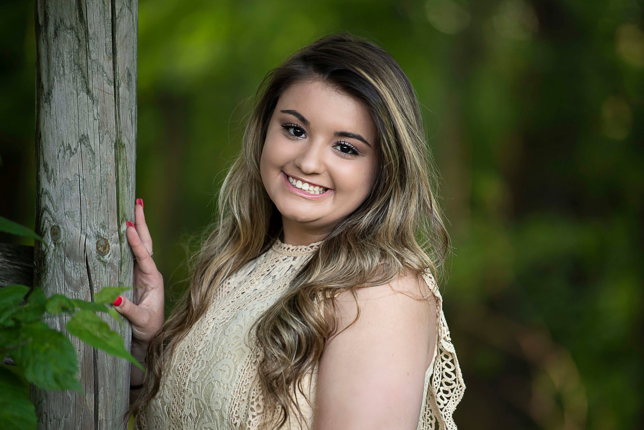 A woman with long, wavy brown hair smiles while leaning against a wooden post. She wears a sleeveless lace top and has painted nails. The background is lush and green, suggesting a forest or garden setting.