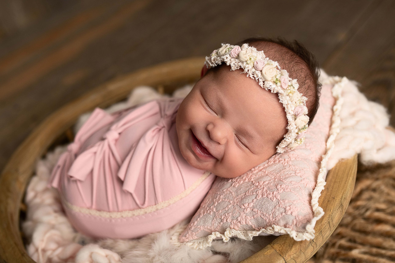A recien nacida, wrapped in a pink blanket, sleeps peacefully in a small wooden frame with a woven net. The baby wears a delicate floral headband and rests on a soft, textured cushion against the wooden floor background.