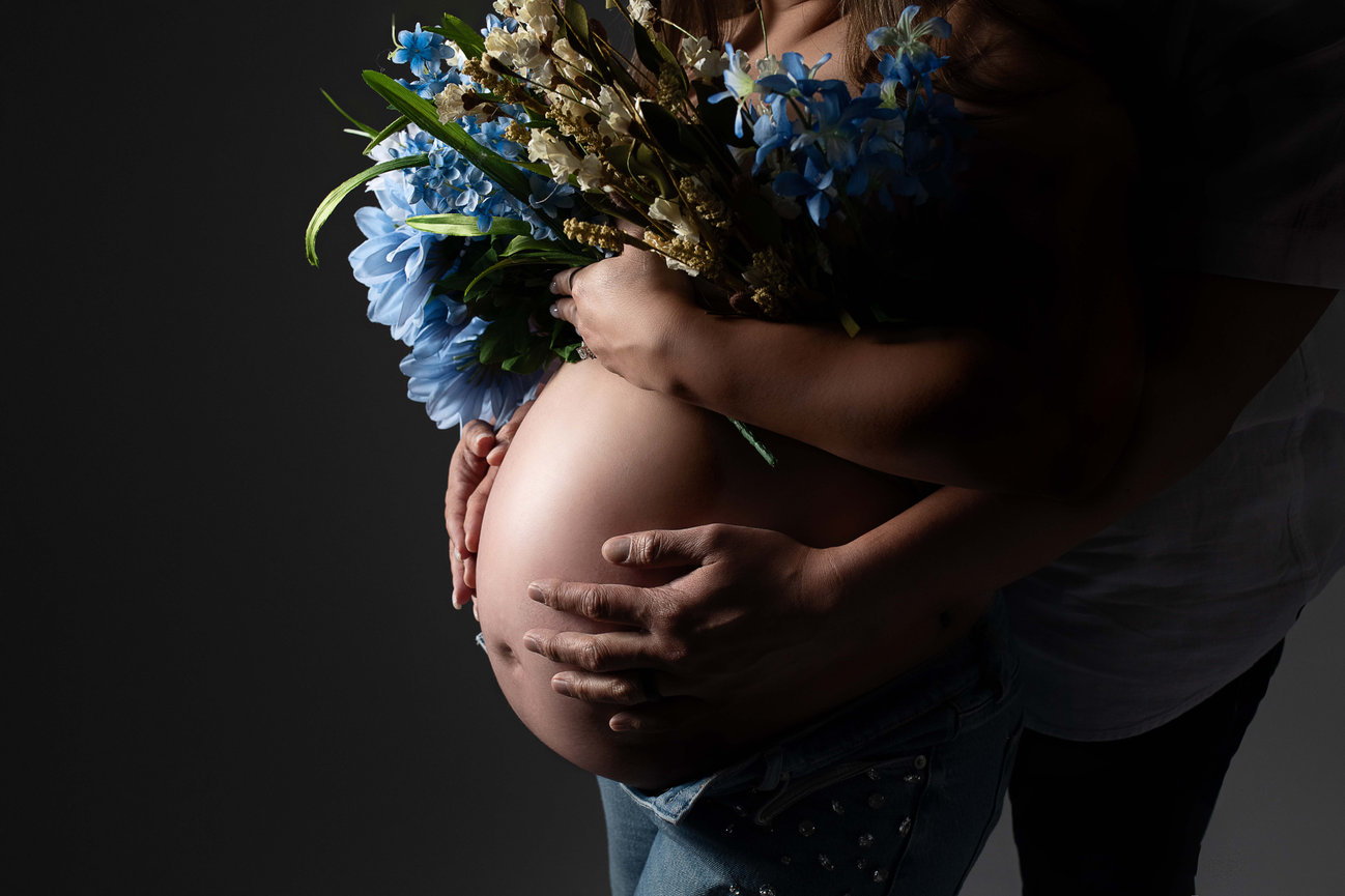 A pregnant person, holds a bouquet of blue flowers against their belly. Another persons hands gently cradle the belly from behind, creating an intimate moment that shines amidst the dark backdrop,  from the bustling suburb of Chicago.