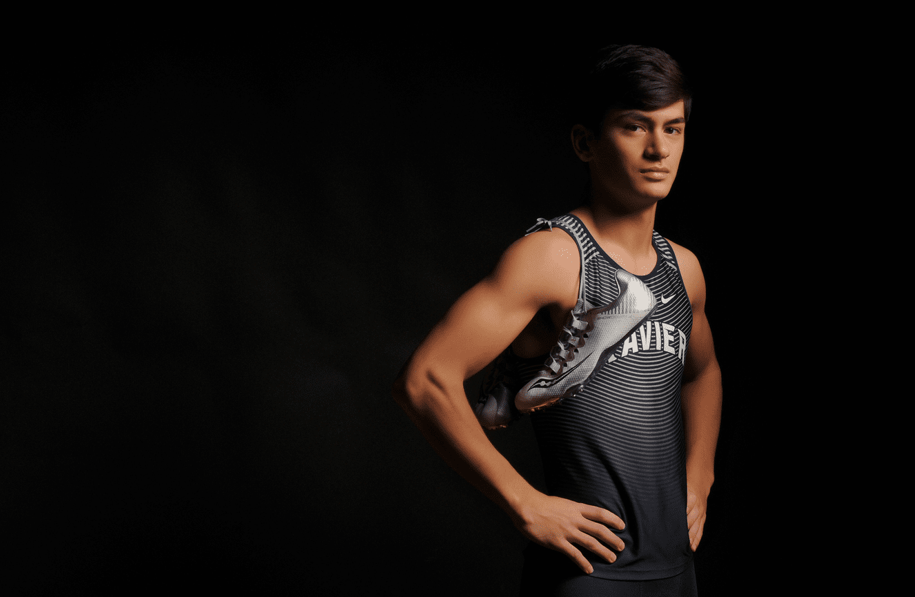 A high school senior track runner poses in his jersey with his running shoes looped over his shoulders.